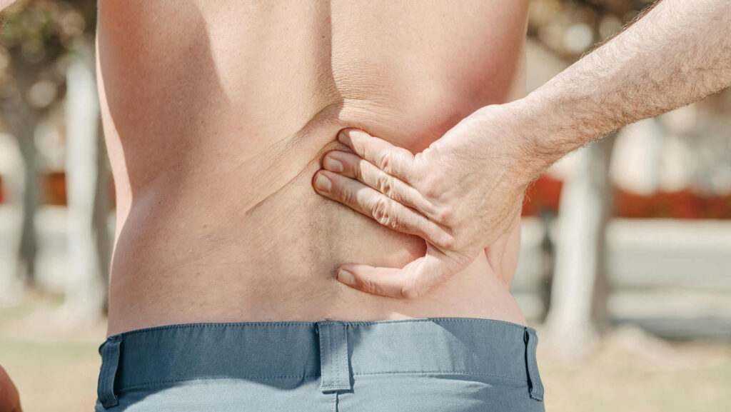 Ein Mann fasst sich mit einer Hand an seinen unteren Rücken, im Bereich der Wirbelsäule.