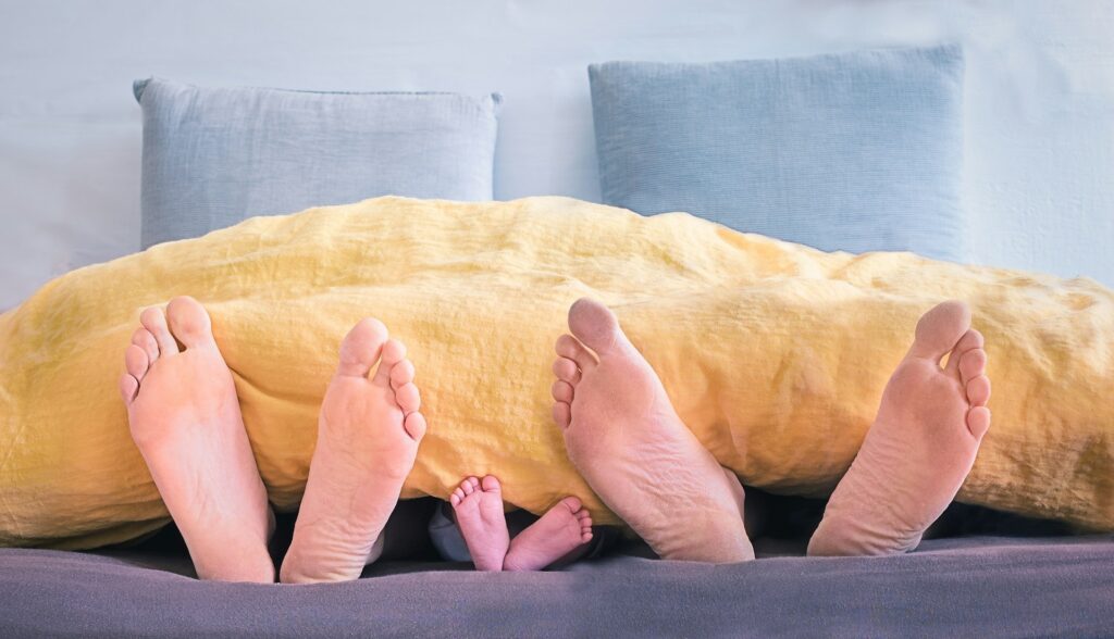 Drei paar nackte Füße, vermutlich eine Familie, sind in einem Bett zu sehen. Hier kann die Rückenlage bei den im Bett liegenden Menschen vermutet werden, welche dabei hilft, die Hüftbeuger im Schlaf zu dehnen