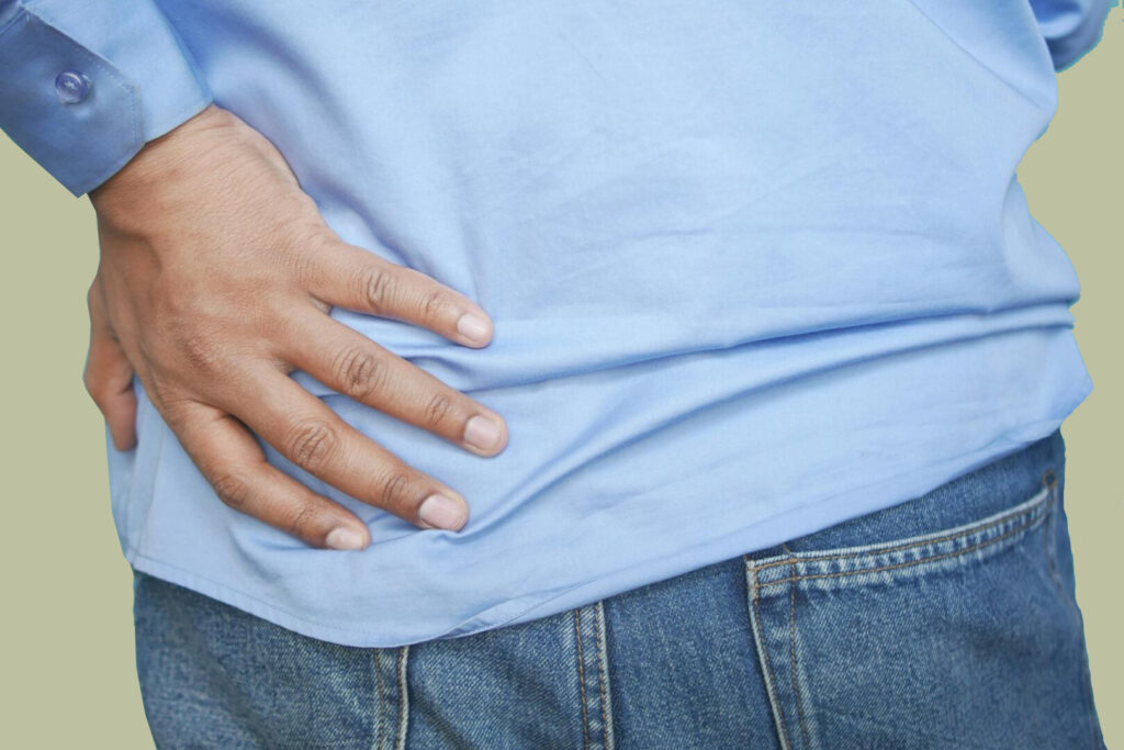 Ein Mann in blauem Hemd ist von hinten zu sehen, der sich mit einer Hand in den Bereich der Lendenwirbelsäule am Rücken greift.