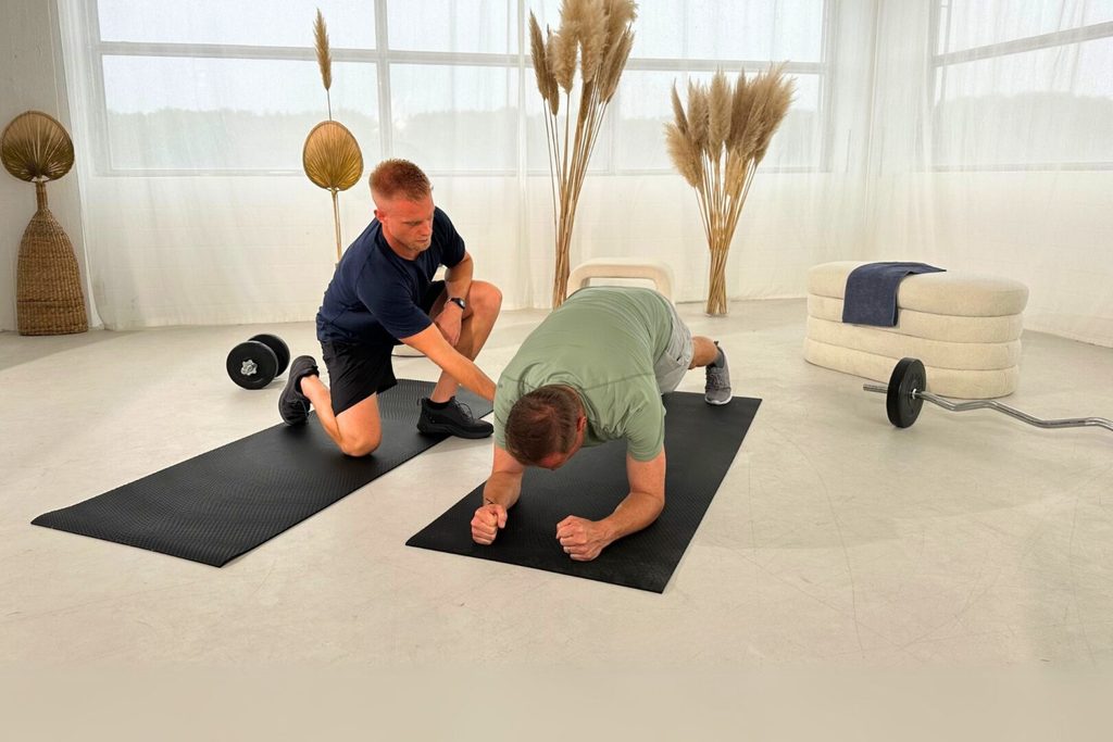 Trainer korrigiert und kommentiert die Plank-Übung eines Mannes beim Präventionskurs, der sich auf den Unterarmen abstützt und eine gerade Körperhaltung hält. Die Übung ist Teil der Wirbelsäulengymnastik